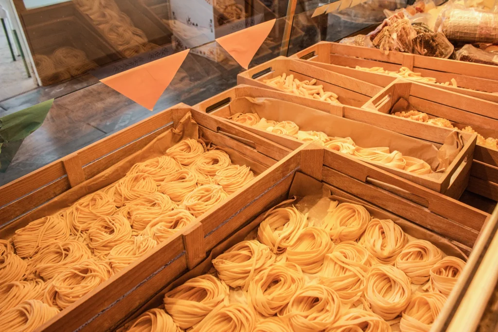 Cajas de madera con pasta fresca tipo tagliatelle o fettuccine, dispuestas en filas detrás de vitrina de cristal; ambiente de tienda gastronómica con productos al fondo.