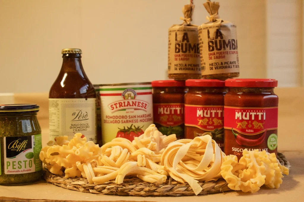 Productos italianos sobre mantel trenzado: pastas crudas tipo tagliatelle y mafaldine, tarros de salsa de tomate Mutti, pesto Biffi, condimentos La Bumba, tomates San Marzano y botella de extracto de orégano.