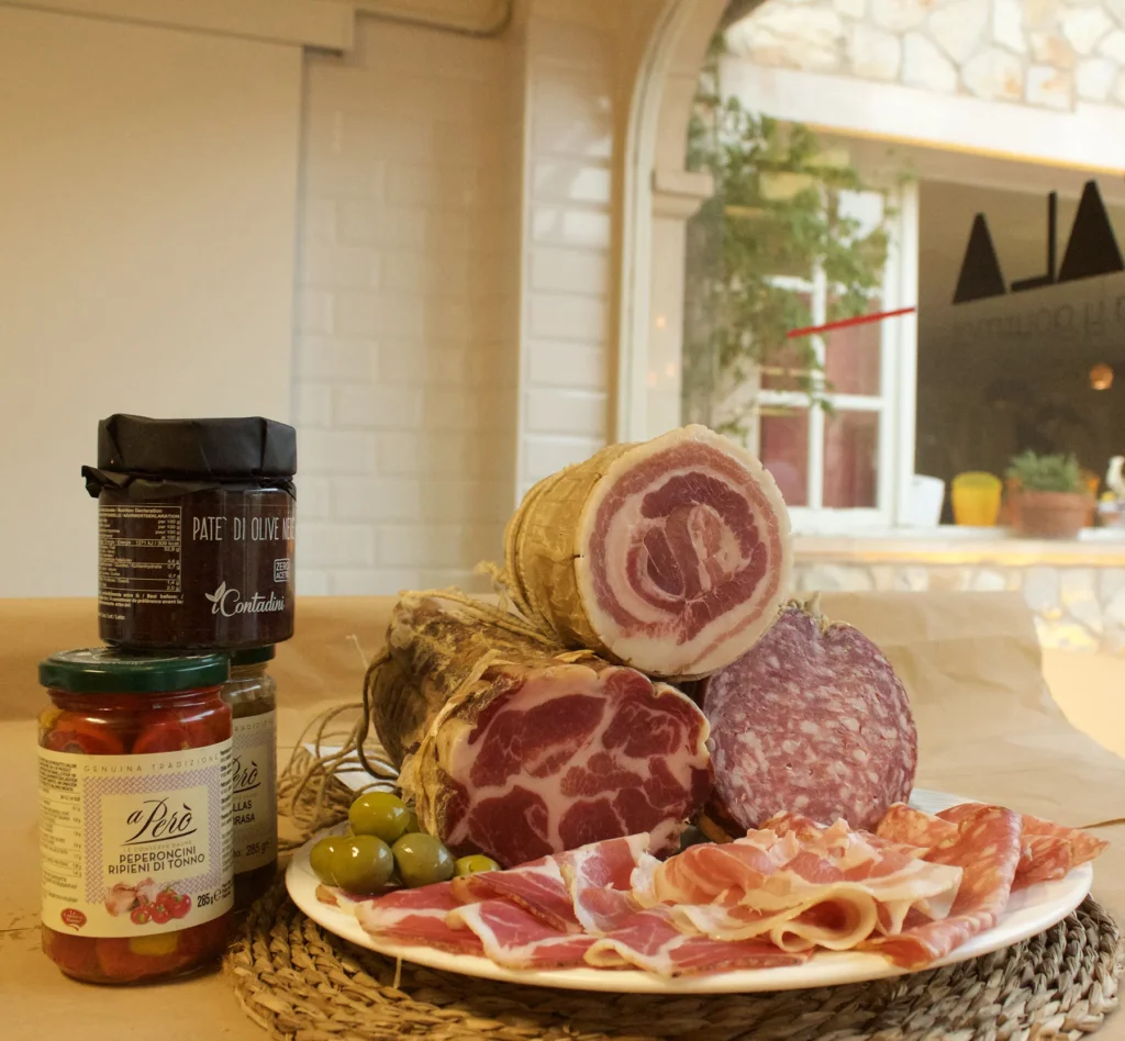 Tabla con embutidos italianos: pancetta enrollada, coppa marmoleada, salami de textura gruesa; plato con lonchas de prosciutto y aceitunas verdes; tarros de condimentos sobre mantel trenzado; fondo con ventana y paisaje mediterráneo.