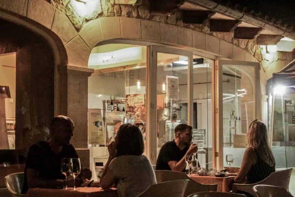 Cena al aire libre en restaurante con fachada de piedra, ambiente cálido y cartel de “BUFALA” visible en el interior.