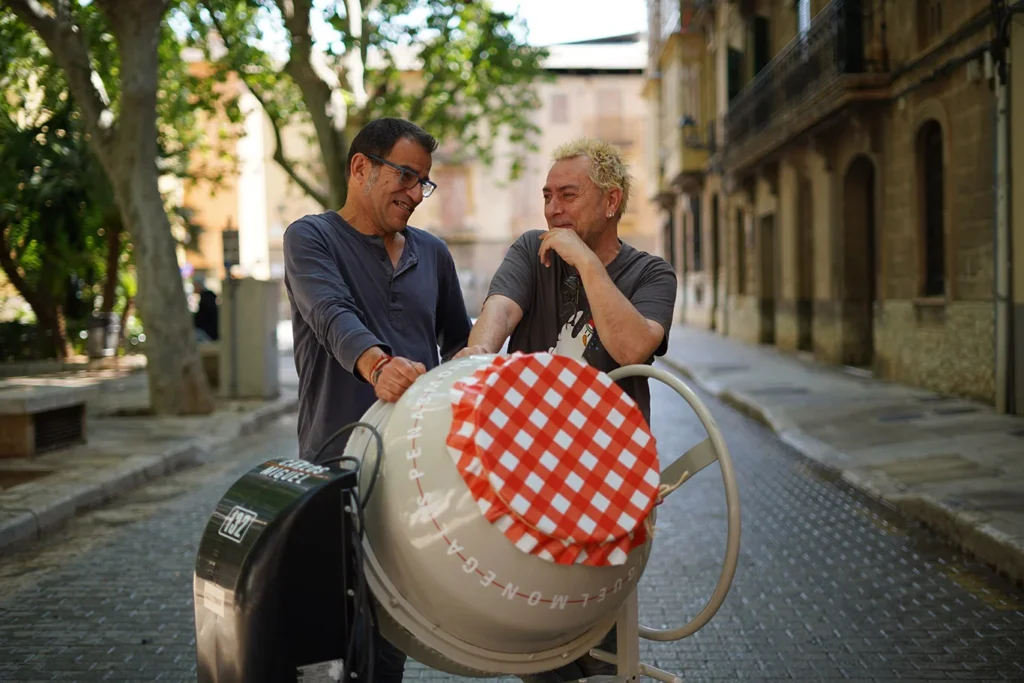 Carlos Penas y Miguel Monegal junto a una hormigonera intervenida en la calle, con fondo urbano y árboles.