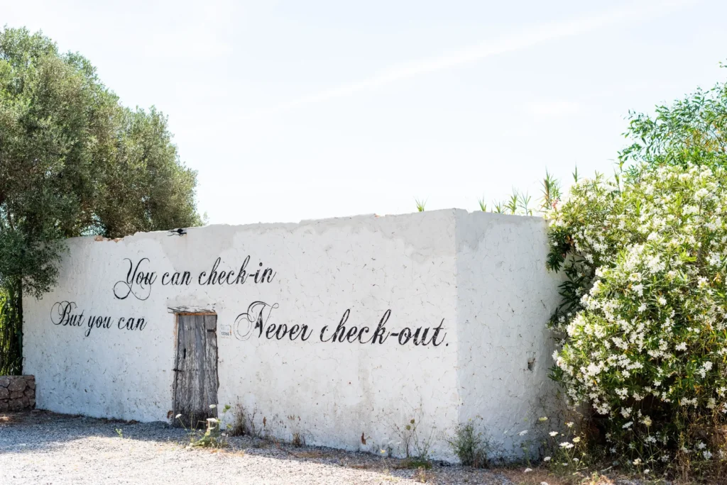 Muro blanco de Pikes Ibiza con puerta de madera y mensaje pintado rodeado de vegetación.