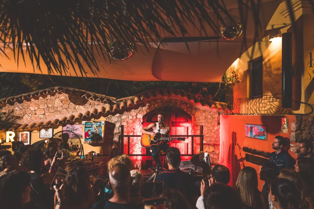 Concierto acústico nocturno en Pikes Ibiza con público alrededor y escenario iluminado en tonos rojos.