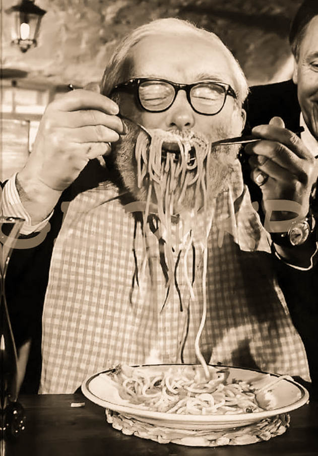Persona mayor comiendo espaguetis con entusiasmo, usando tenedor y cuchara; gran cantidad de pasta colgando de la boca; ambiente cálido con otra persona sonriendo al fondo.