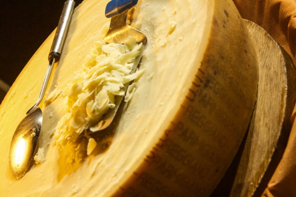 Rueda de queso Parmigiano-Reggiano parcialmente rallada con rizos finos sobre una superficie de madera; espátula y cuchara metálica colocadas encima; paño naranja visible al lateral.