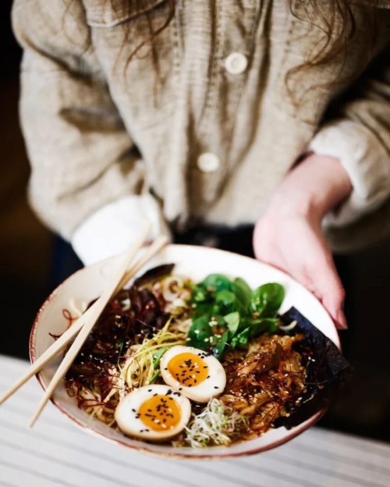 Bowl de ramen con huevos marinados, verduras frescas y fideos sostenido entre las manos.