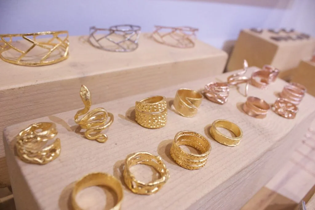 Exposición de anillos y pulseras artesanales en plata, oro y oro rosa sobre soportes de madera.