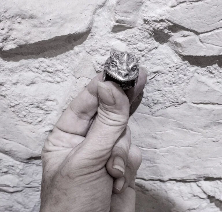 Anillo artesanal con cabeza de criatura reptil tallada, sostenido en la mano.