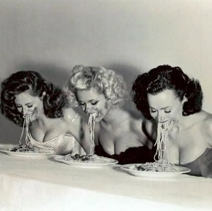 Tres personas comiendo espaguetis en sincronía, vestidas con estilo retro, en fotografía en blanco y negro.