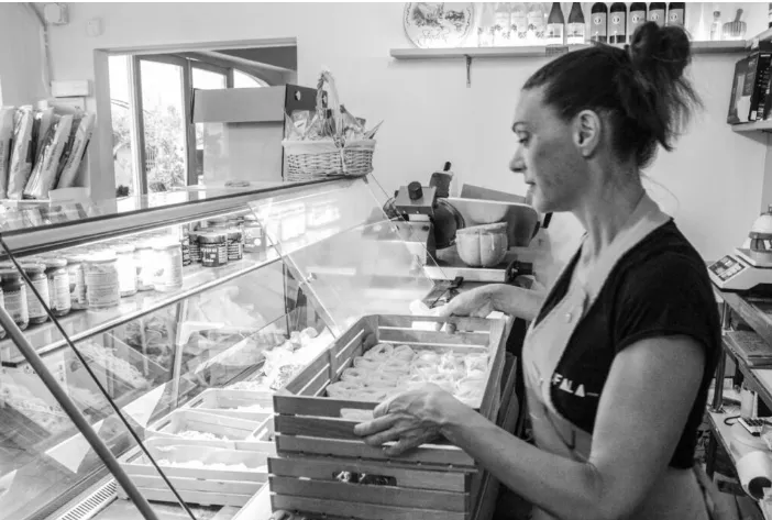 Mujer tras mostrador de tienda local con vitrina de productos, bandeja de bollería y estanterías con conservas y utensilios.