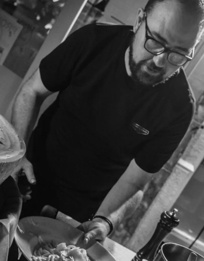 Persona sirviendo pasta sobre mostrador de cocina con molinillo de pimienta y cuenco grande en primer plano.