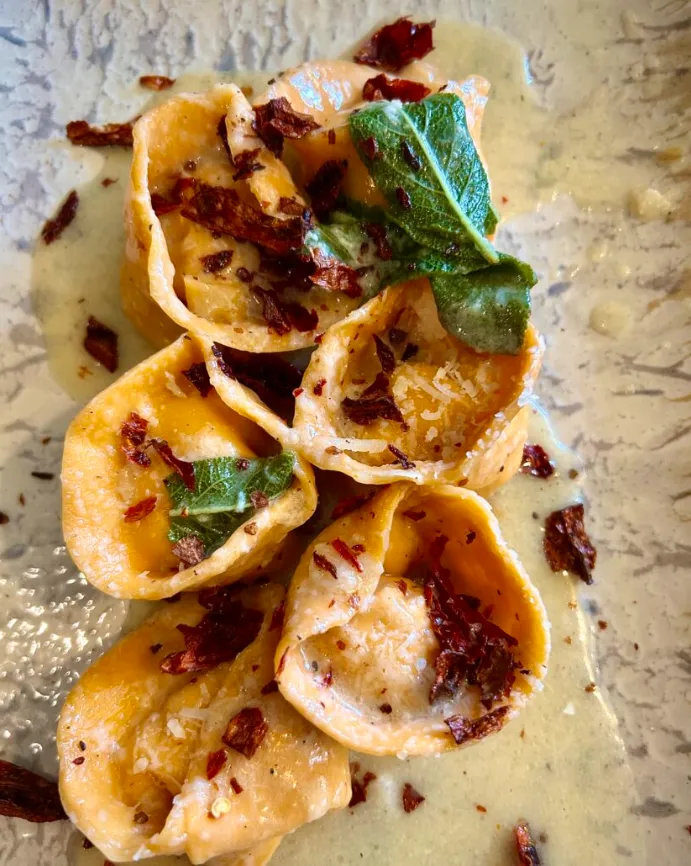 Tortellini en salsa cremosa con hojas de salvia y crujientes rojos sobre plato texturizado.