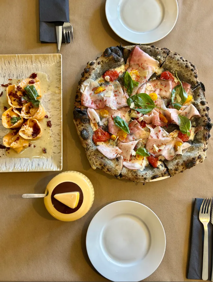 Mesa rústica con pizza de jamón y albahaca, pasta rellena con salsa de tomate y vajilla dispuesta para dos comensales.