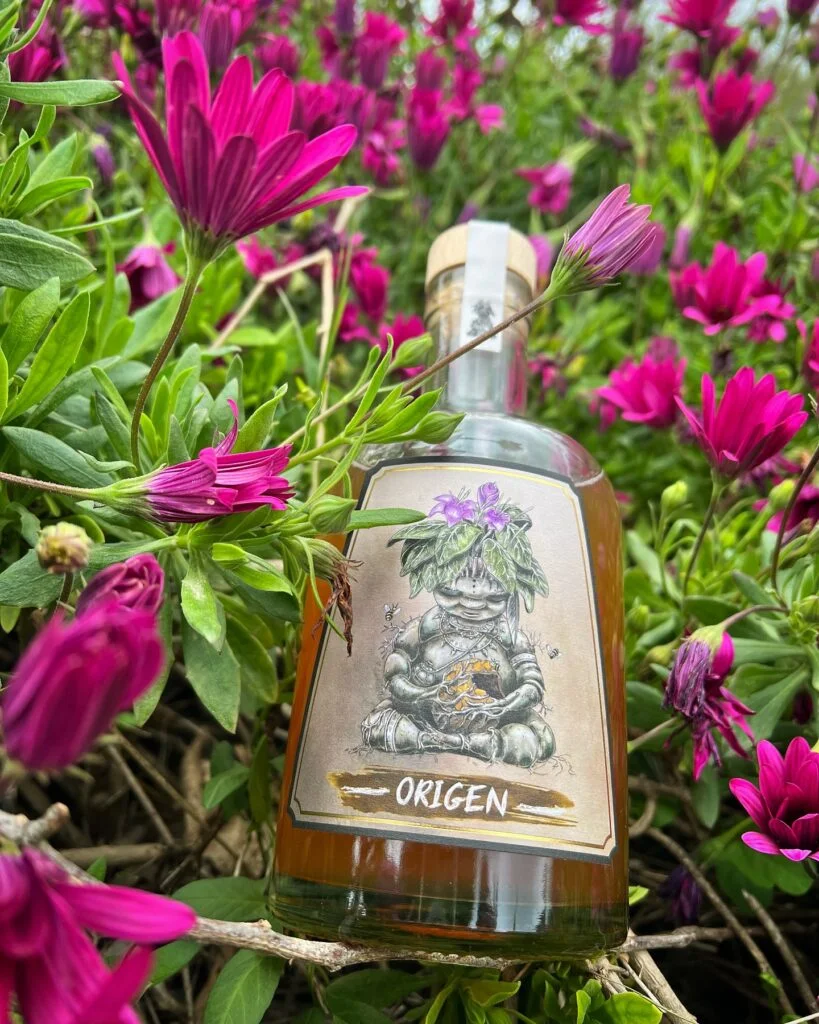 Botella de Mandrágora Hidromel Dulce entre flores moradas y follaje verde, con etiqueta artesanal de estilo heráldico.