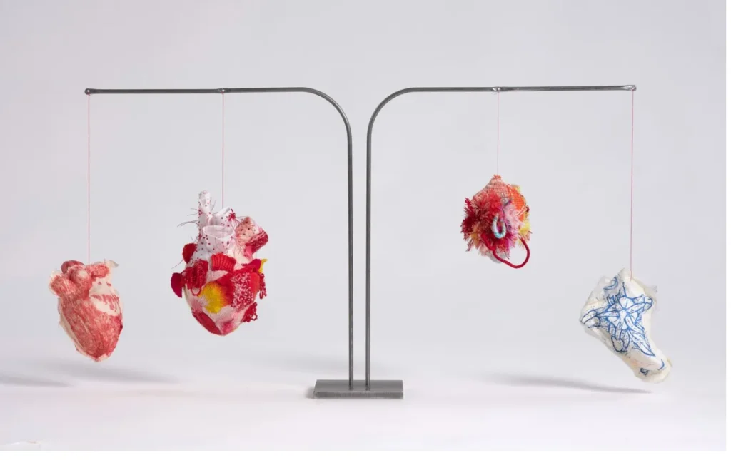 Instalación artística con cuatro corazones anatómicos suspendidos, elaborados con textiles coloridos.