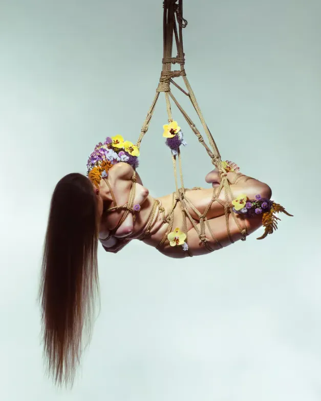 Figura suspendida en el aire mediante cuerdas decorativas, adornada con flores de colores sobre fondo azul etéreo.