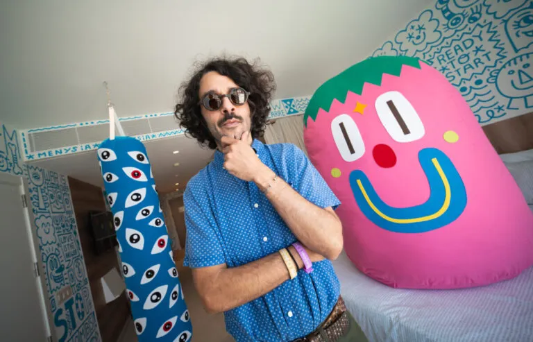 El artista Albert Pinya con camisa azul de lunares posa pensativo en una sala decorada con arte pop y figuras de peluche gigantes.