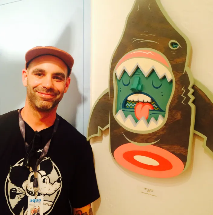 Alex Yanes junto a su obra “Sharkface”, escultura mural de tiburón con rostro caricaturesco y expresión angustiada