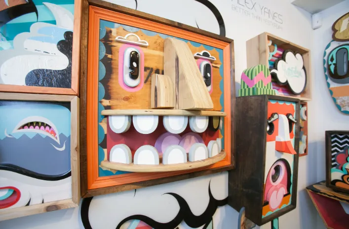Instalación mural de Alex Yanes con esculturas de rostros caricaturescos en madera pintada, fondo con diseño gráfico y lema “Better Than Yesterday”