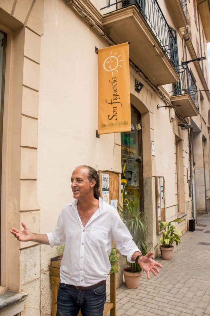 Carlos Salas con camisa blanca frente a la herbodietética ecológica Son Bugadella en Inca, con fachada beige, plantas en la entrada y cartel con sol sonriente.