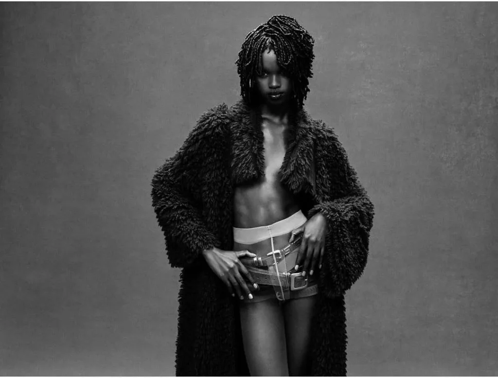 Persona con abrigo oscuro texturizado, shorts vaqueros y peinado trenzado; retrato en blanco y negro con pose segura y estética artística.