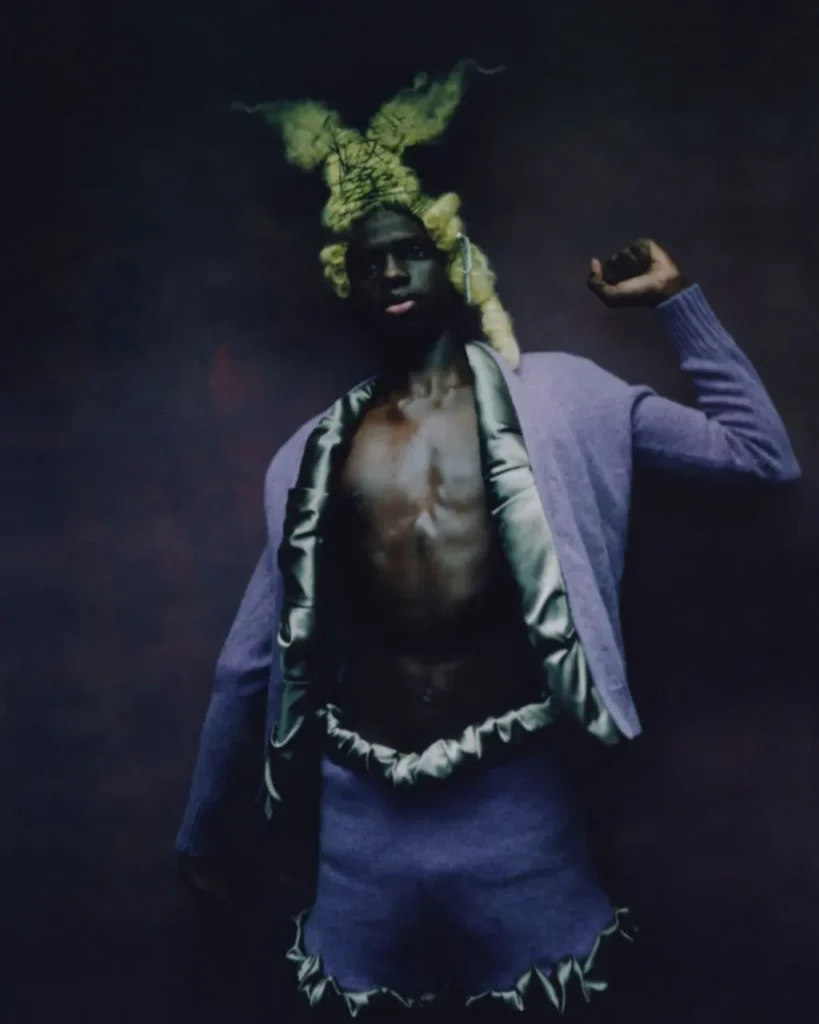 Persona con conjunto lavanda y tocado verde en forma de cuernos; retrato artístico con fondo oscuro y estética avant-garde.