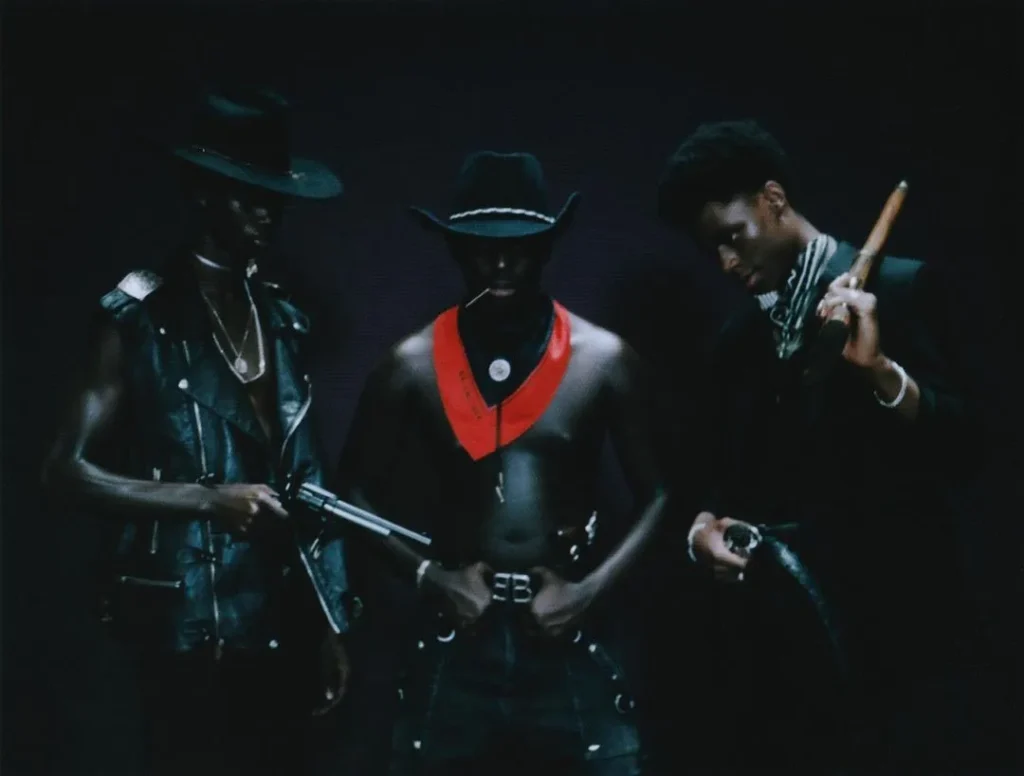 Tres personas con atuendos western estilizados, sombreros, armas y bate; retrato grupal en fondo oscuro con actitud escénica y moda narrativa.