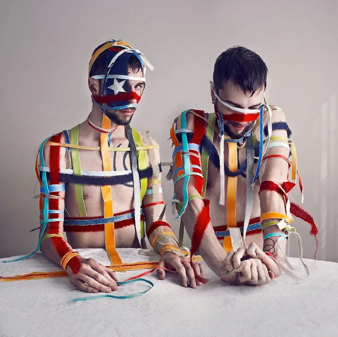 Dos figuras sentadas con el rostro y cuerpo envueltos en tiras de tela multicolor, formando patrones abstractos que evocan banderas y estructuras sociales.