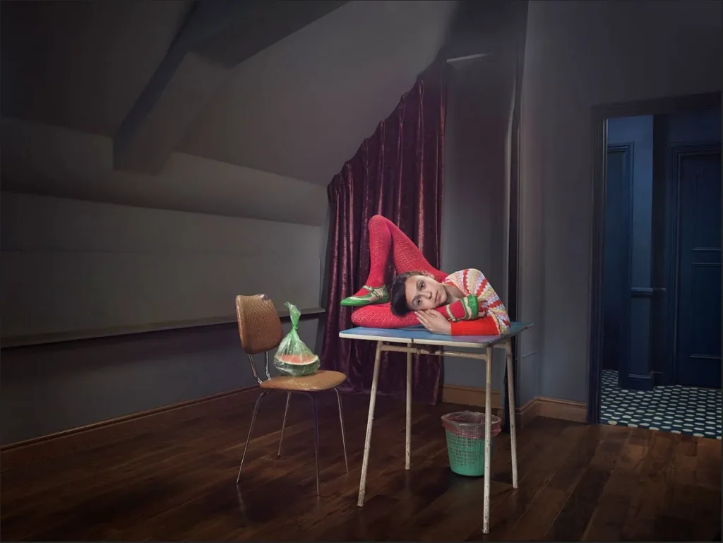 Persona en pose contorsionista sobre mesa en habitación oscura con cortina roja
