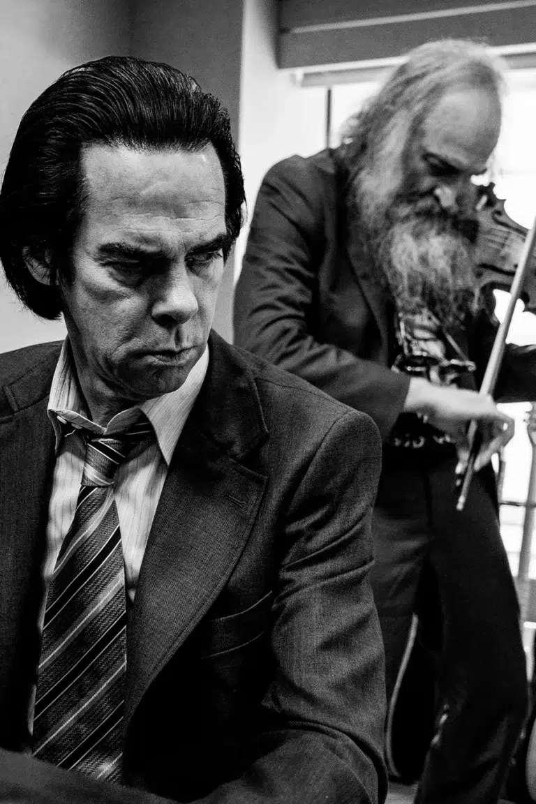Nick Cave con expresión introspectiva en primer plano, mientras Warren Ellis toca el violín al fondo en una fotografía en blanco y negro.