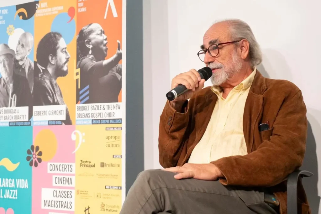 Roberto Menéndez habla al micrófono durante una entrevista en Palma. A su lado, un cartel colorido anuncia eventos culturales.