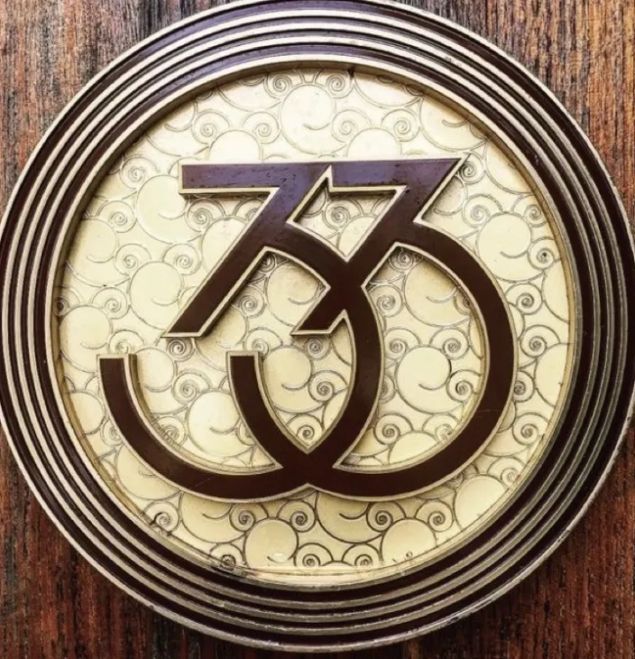 Emblema circular con diseño ornamental que entrelaza los números 73 y 33 sobre fondo beige con motivos ondulados.