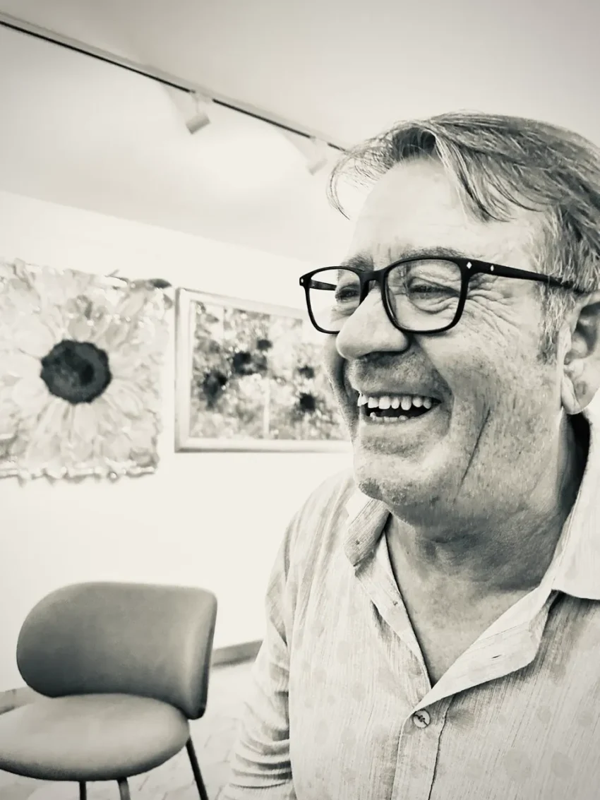 Retrato en blanco y negro de persona mayor con gafas y camisa clara estampada, sonriendo en una sala decorada con arte floral y mobiliario moderno.