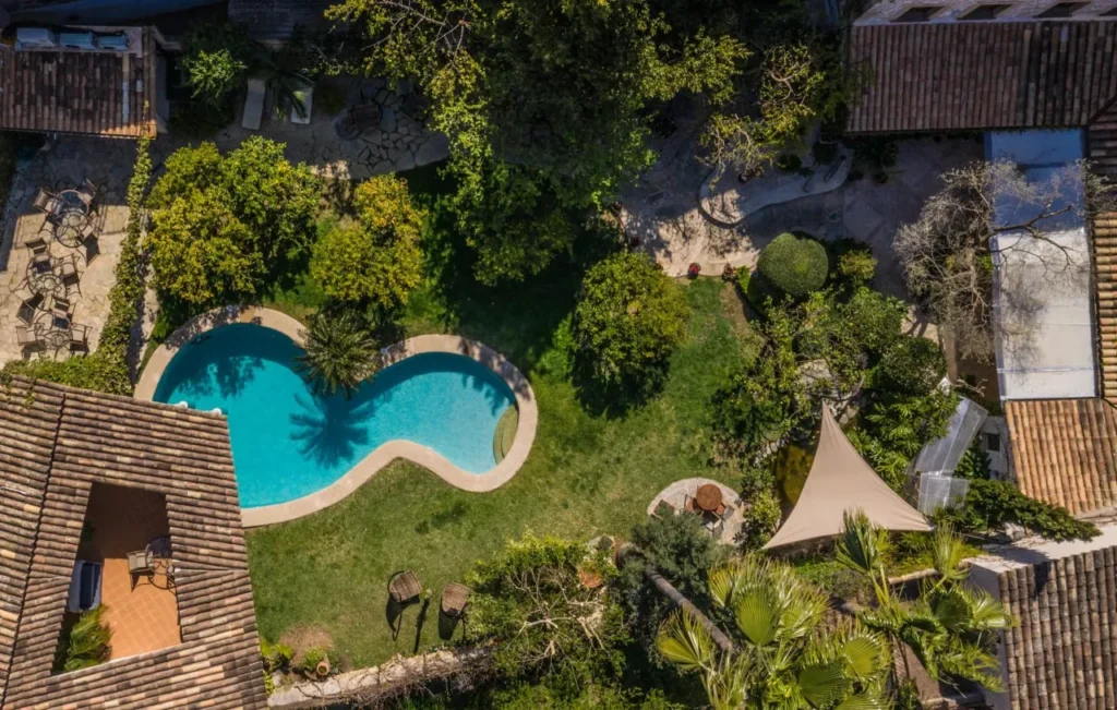 Vista aérea de jardín con piscina en forma de riñón, rodeada de césped, árboles y edificios con tejados de teja en Mallorca.
