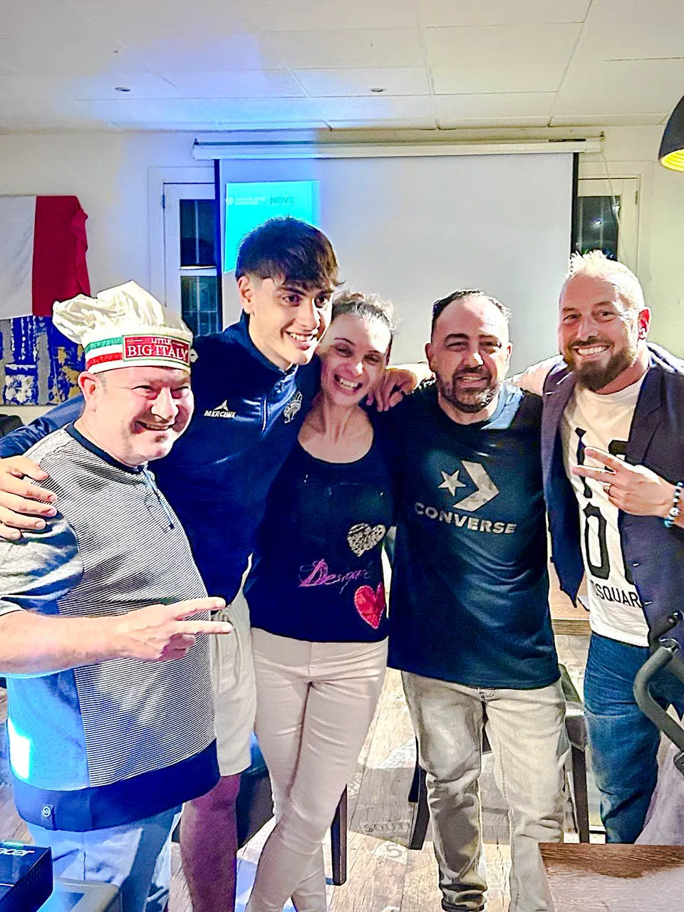 Cinco personas sonrientes en un evento festivo, una con gorro de chef “BICHYAY”, fondo con bandera francesa y proyector
