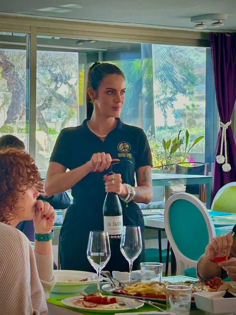 Camarera sirviendo vino La Bastide en restaurante Moka Piu de Palma, junto a mesa con comensales en conversación, decoración moderna y vista exterior.