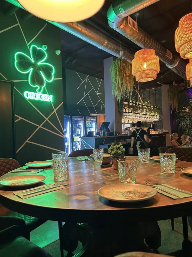 Interior de restaurante moderno en Palma con mesa redonda, lámparas tejidas, barra iluminada y letrero de neón “ORIGEN” con trébol.