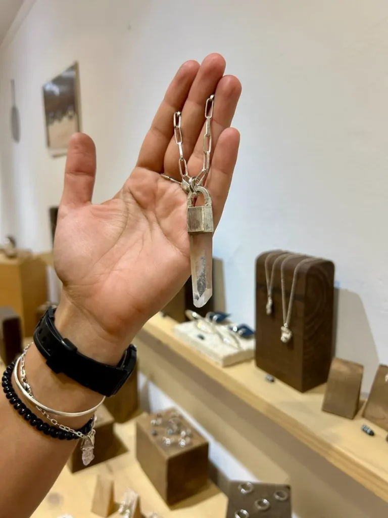 Mano sosteniendo un colgante de cuarzo transparente con cadena de eslabones plateados, frente a un expositor de joyas artesanales en una boutique de Palma.