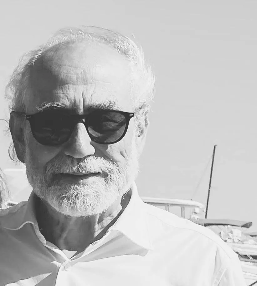 Retrato en blanco y negro de Roberto Menéndez con gafas de sol en un puerto, rodeado de mástiles y barcos.
