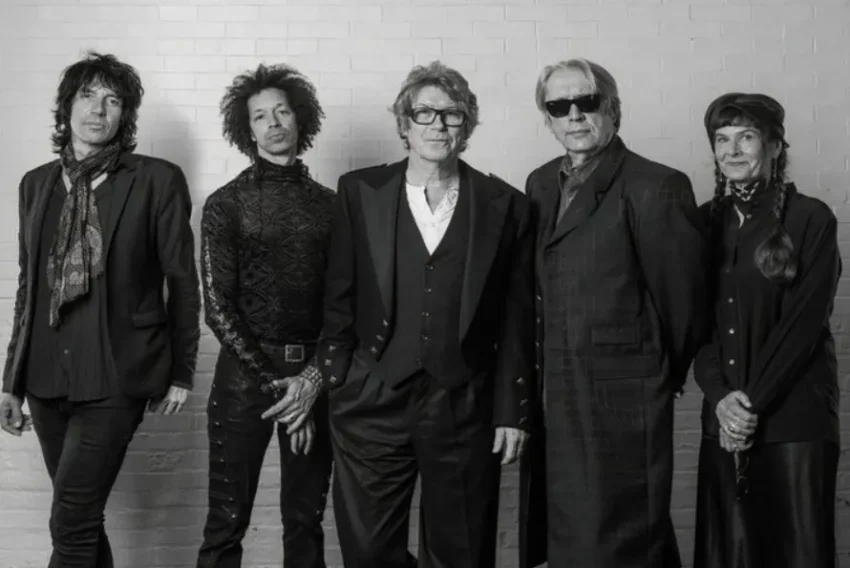 The Psychedelic Furs vestidos con ropa oscura y estilizada posan frente a una pared de ladrillo claro; su actitud y vestimenta sugieren una identidad artística o performativa.