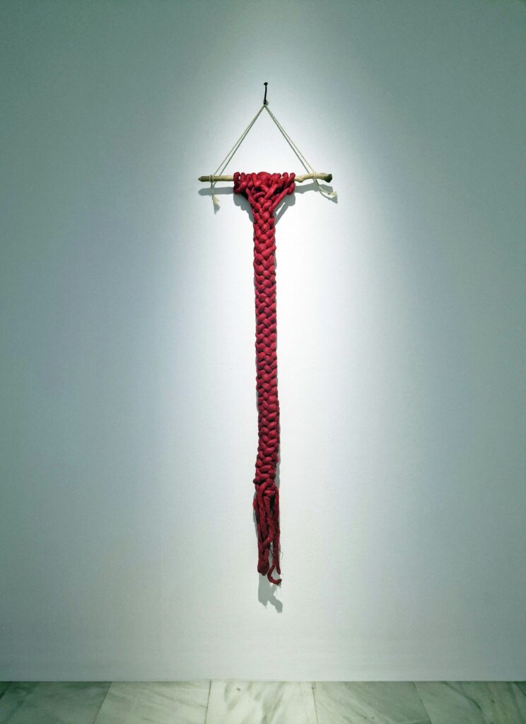 Escultura de Carlos Nicanor de una trenza de algo más de un metro elaborada con cuerda roja gruesa que cuelga en la pared sujeta por un tronco en el principio