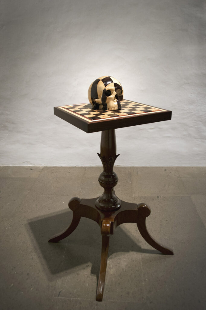 La escultura de Carlos Nicanor representa una mesa de ajedrez de madera, con un tablero con los típicos cuadrados blanco y negro (en el caso tonalidades de  la misma madera) y en vez de las figuritas una clavera encima siguiendo el patrón  de los cuadrados 