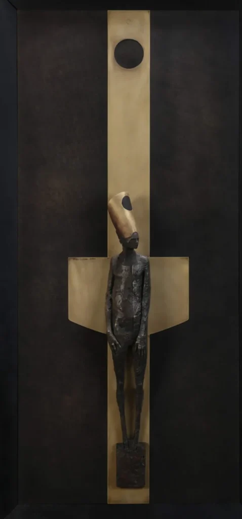 Figura humana estilizada con tocado cónico frente a cruz dorada, escultura de Max Leiva