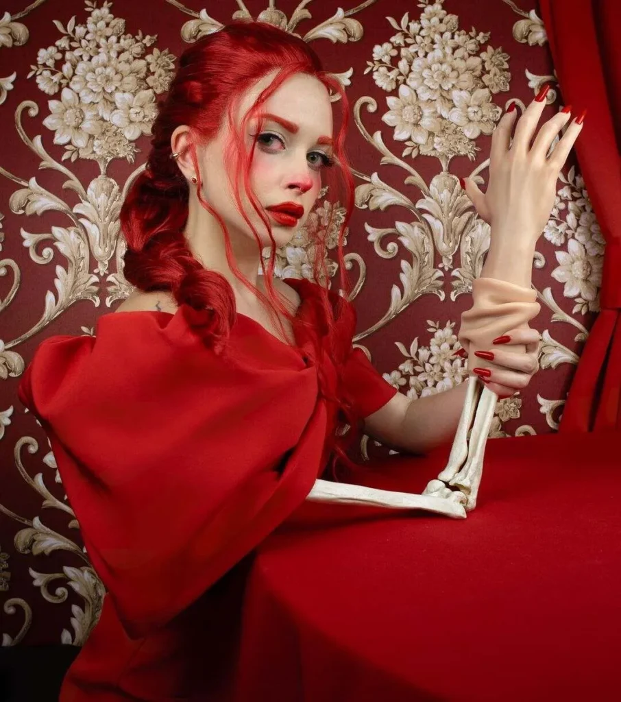 Persona pelirroja con trenzas sostiene un brazo esquelético cubierto parcialmente por un guante, frente a fondo rojo floral.