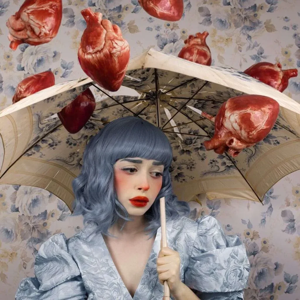 Persona con cabello azul grisáceo sostiene paraguas floral mientras corazones anatómicos llueven sobre fondo vintage.