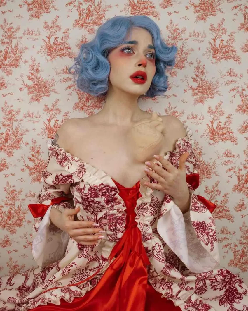 Persona con cabello azul rizado y maquillaje dramático sostiene un busto escultórico, vestida con atuendo aristocrático frente a fondo rojo estampado.