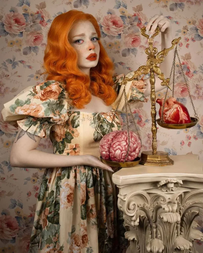 Mujer pelirroja con vestido floral sostiene una balanza dorada con un cerebro en un lado y un corazón en el otro, frente a fondo de papel pintado floral.