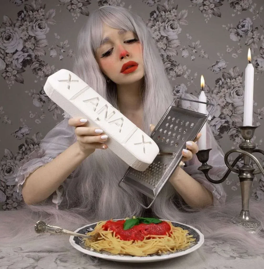 Mujer con cabello plateado y maquillaje dramático ralla una pastilla gigante de Xanax sobre un plato de espaguetis en un entorno surrealista con candelabro y papel pintado floral.
