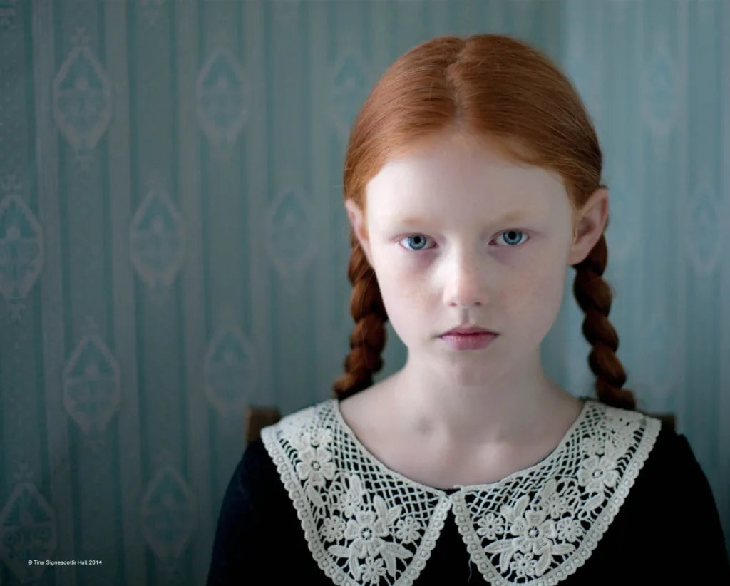 Retrato clásico de joven pelirroja con trenzas, vestido negro y cuello de encaje blanco, fondo azul simétrico.