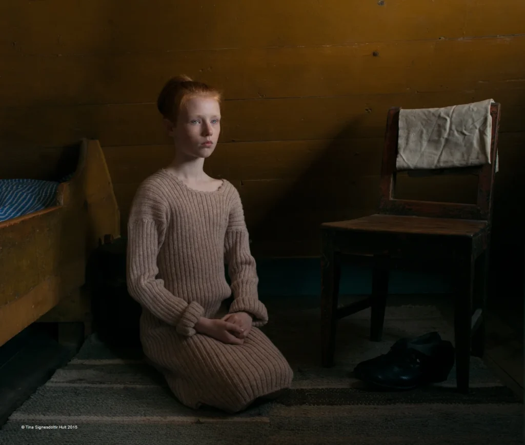 Joven pelirroja arrodillada en habitación rústica, entre cuna de madera y silla con tela blanca.